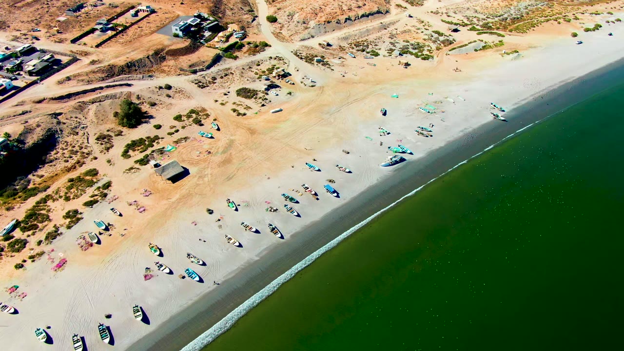 San Juanico aerial frame with green ocean occupying the right side, a bright surf edge, several small boats on the sand, arid dune terrain above, and no visible people.