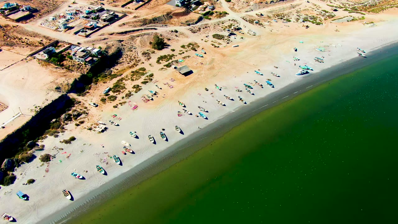 Opening aerial frame over San Juanico, Baja California Sur, showing small boats scattered on pale sand beside green shoreline water and low buildings inland.
