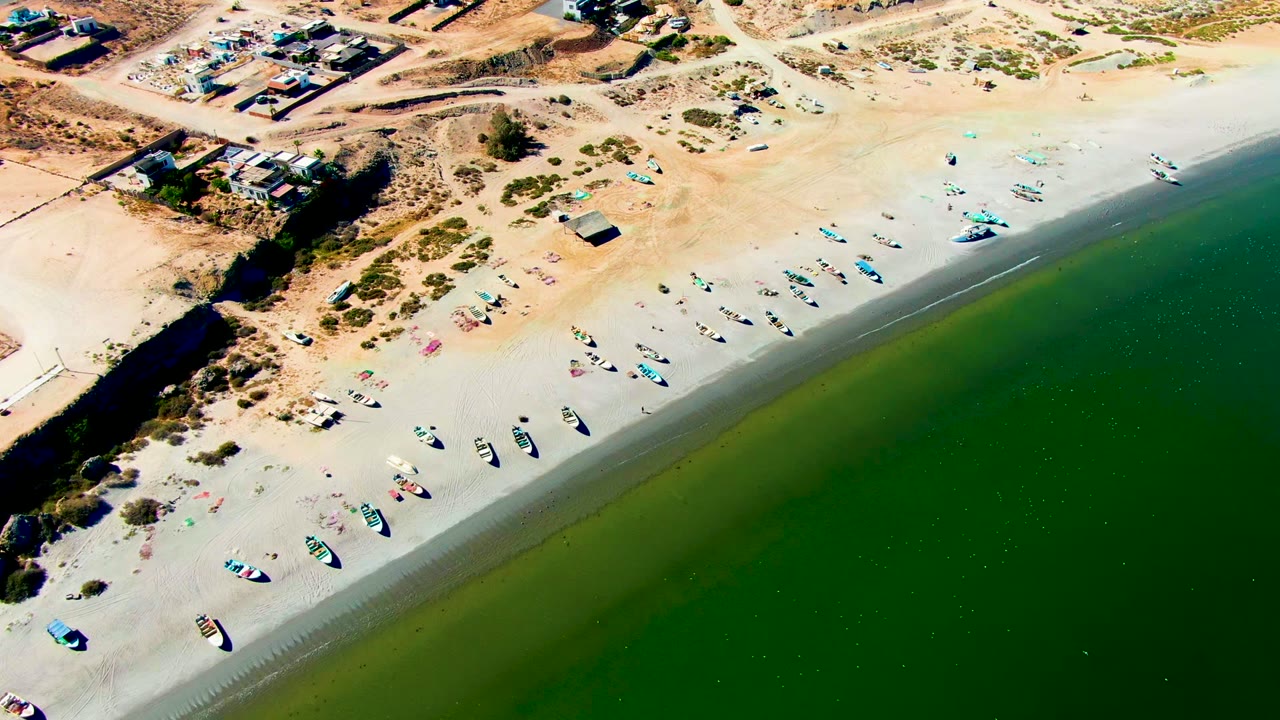 Drone view of boats lined across a sandy beach beside green ocean water.