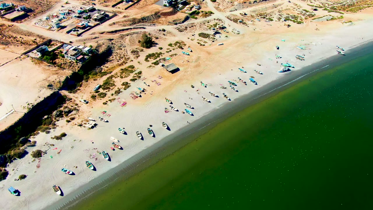 San Juanico aerial frame with the camera looking down across inland buildings, sparse brush, pale open sand, clustered boats, and green water along the right edge.