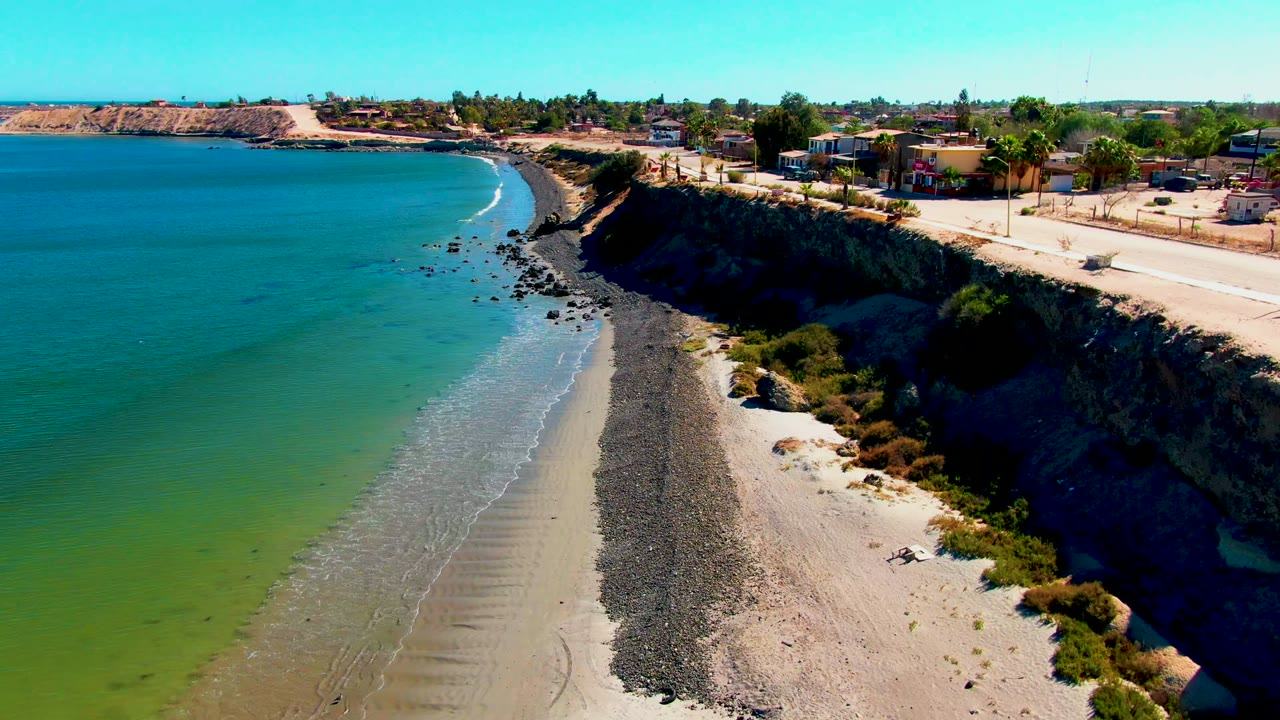 Early frame of San Juanico, Baja California Sur with blue-green ocean at left, a narrow pebbled beach below the cliff, road and homes above, and no visible people.