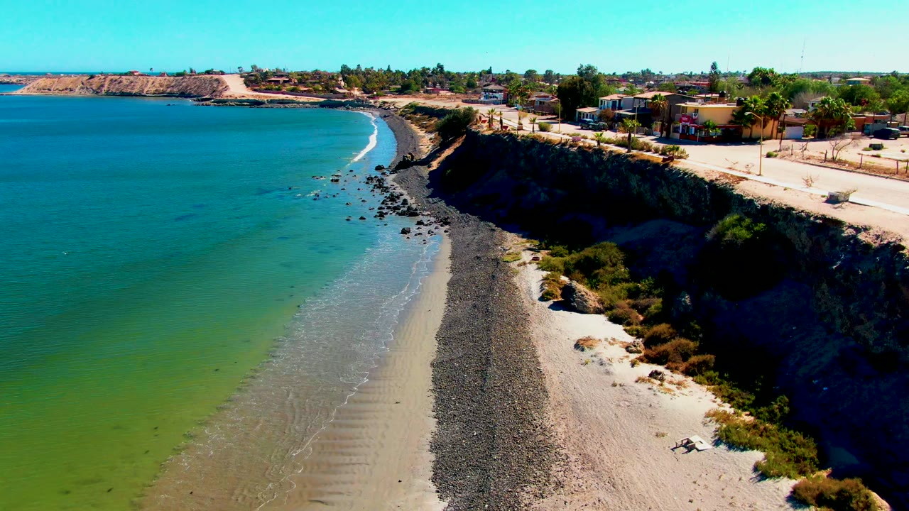 Drone view along a curving beach beneath a cliffside road and houses.