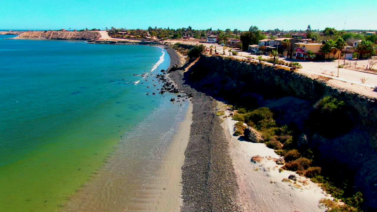 Mid-early San Juanico frame with more open blue water visible, surf tracing the pebbled beach, cliffside road and scattered homes above, and no visible people.