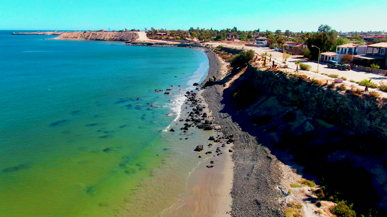 Aerial frame over San Juanico, Baja California Sur, showing blue-green ocean, a curved rocky beach, dark cliff edge, coastal road, low buildings, and tiny people near the roadside.