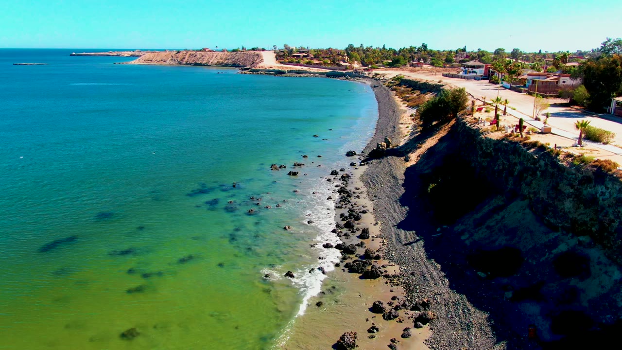 San Juanico aerial frame with the ocean filling the left side, a narrow rocky beach running down the center, a shadowed cliff on the right, and small roadside structures above.