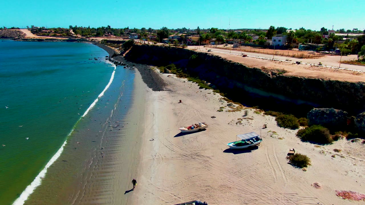 Aerial frame over San Juanico, Baja California Sur shows green-blue water washing onto sand, two beached boats near the lower shore, a cliff with road above, low buildings in back, and a small person visible on the beach.