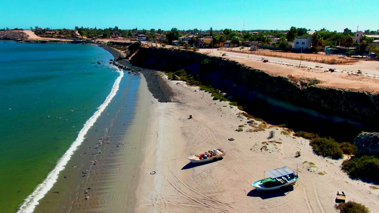 Wide drone view of waves reaching a beach with two boats on sand and a cliff road behind.