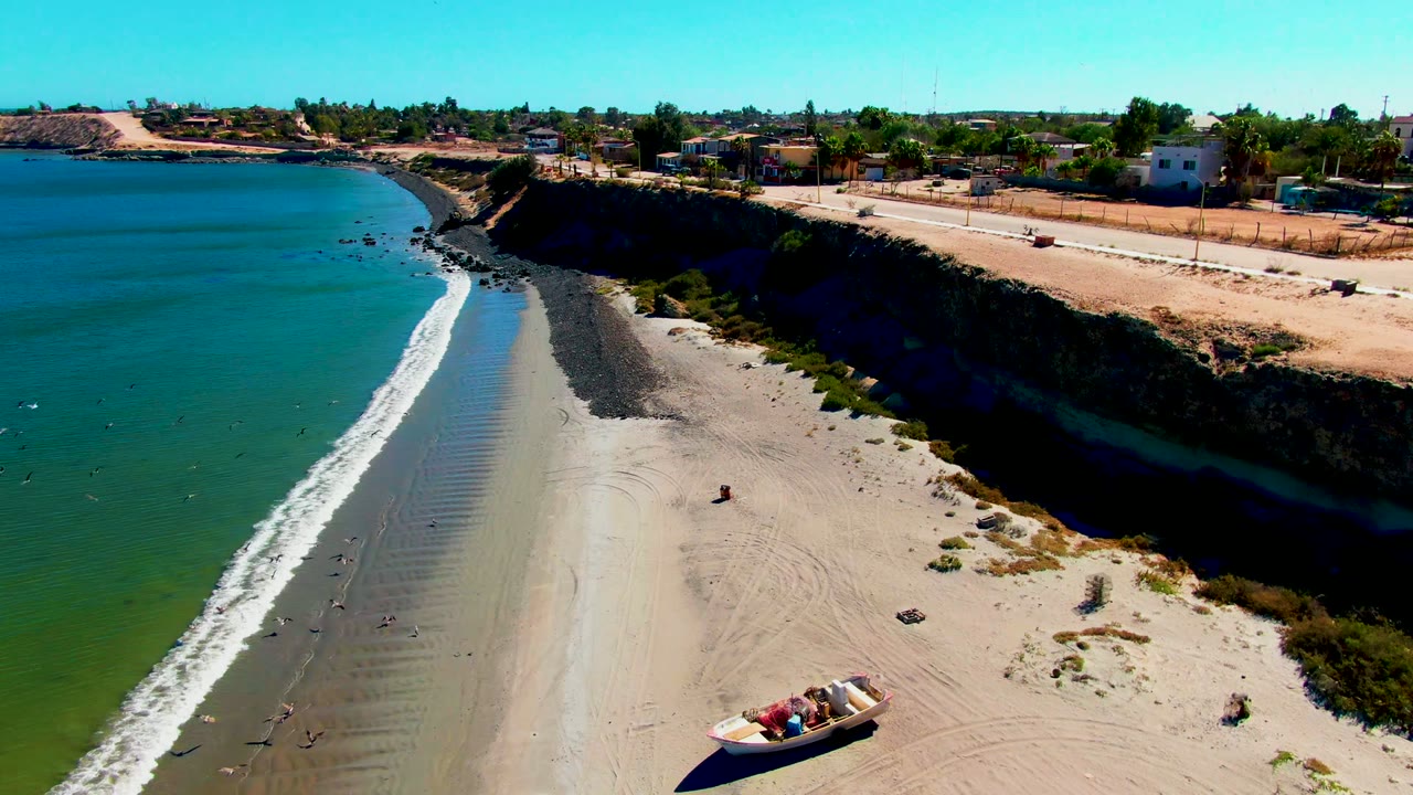 At 0:02, the aerial view shifts along the San Juanico shoreline, placing the beached boats lower in frame while the curved surf line, rocky dark beach band, cliff road, and low buildings stay visible.