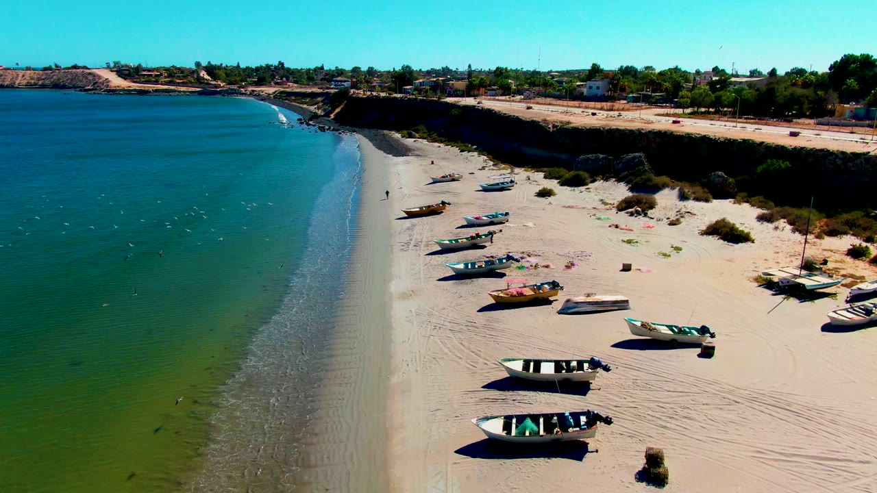 Aerial frame over San Juanico, Baja California Sur, showing multiple small boats on pale sand beside green-blue water, with a bluff and distant buildings beyond the beach.