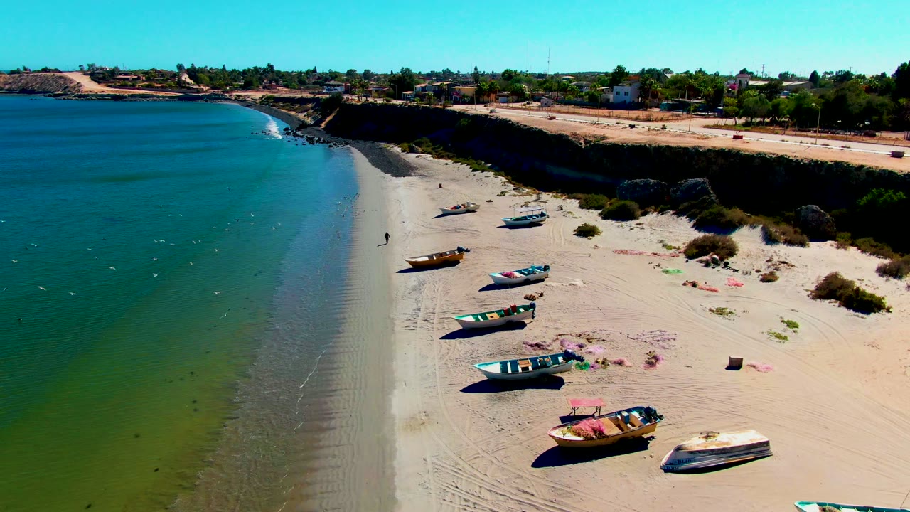 San Juanico beach from above, with fewer visible boats on the sand, shallow water on the left, and low coastal vegetation and buildings beyond the bluff.