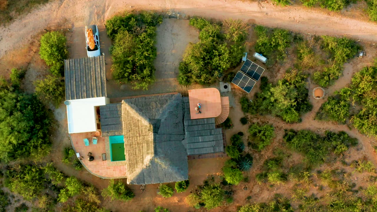 Overhead frame from Cabo Pulmo, Baja California Sur showing a car near the top dirt road, a thatched-roof house below it, solar panels to the right, dense trees, and no visible people.