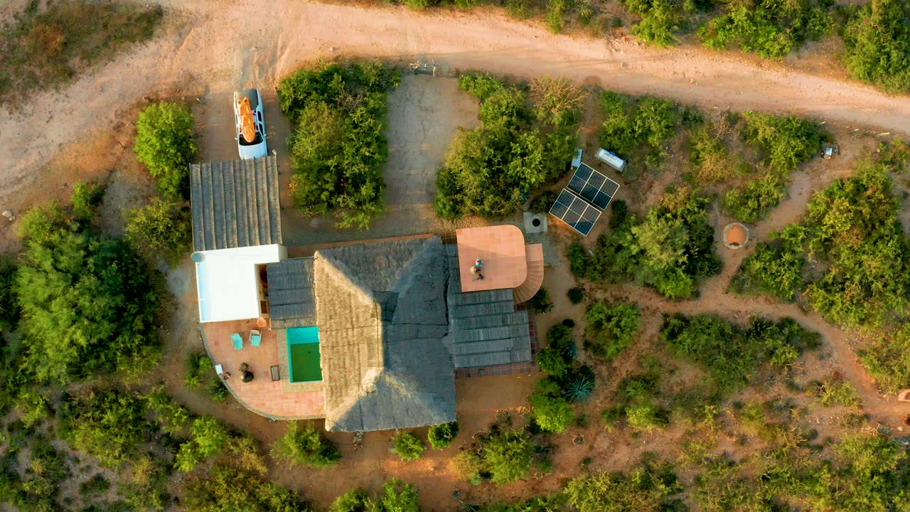 At 0:02, the car remains on the pale dirt road near the top of frame, with the house roof, patio, solar panels, trees, and dry ground visible below in Cabo Pulmo, no visible people.