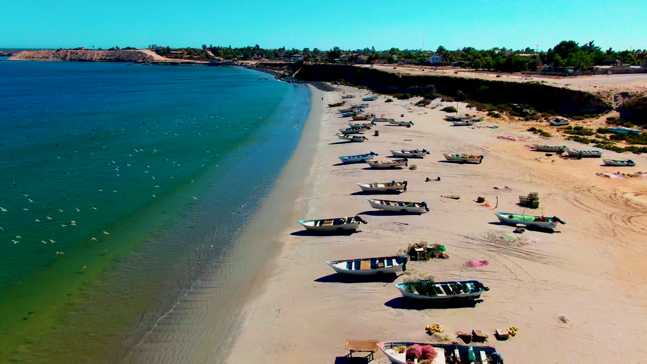 Aerial frame from San Juanico, Baja California Sur showing fishing boats scattered across pale sand, blue-green ocean on the left, and distant low buildings beyond the beach.