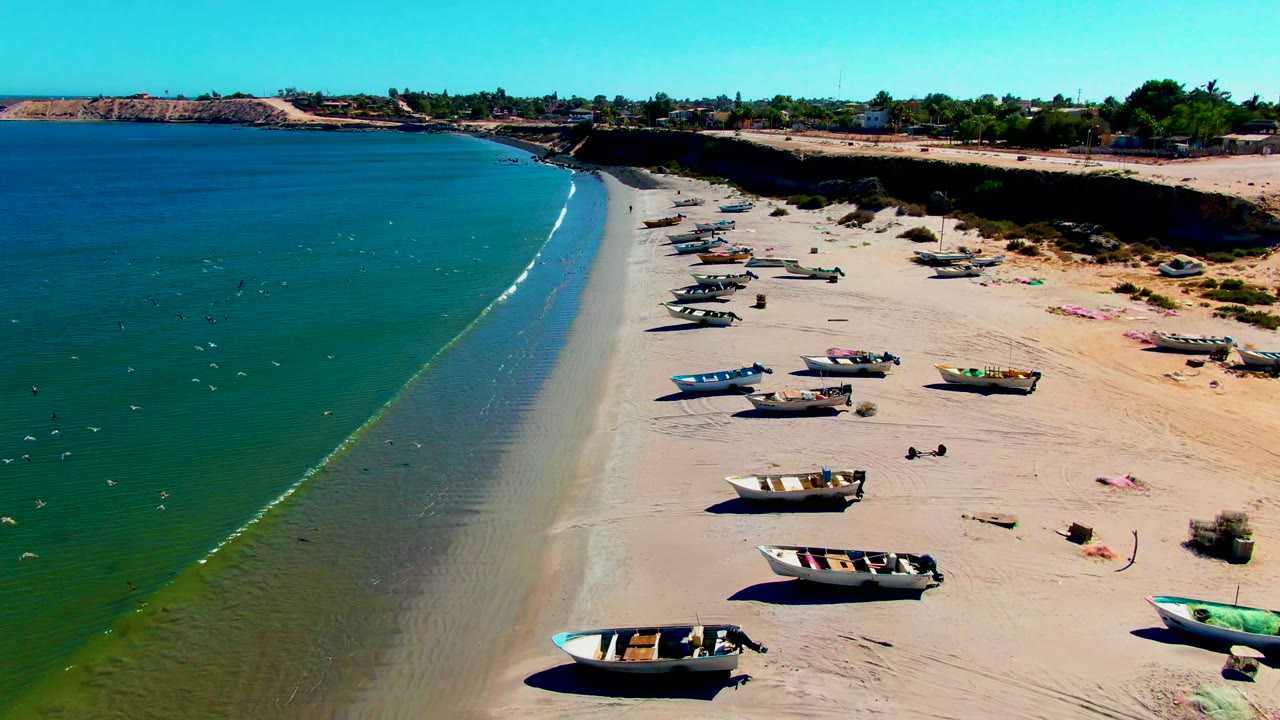 Aerial frame from San Juanico, Baja California Sur where a brighter surf line begins to form along the beach, with boats placed in rows across the sand.