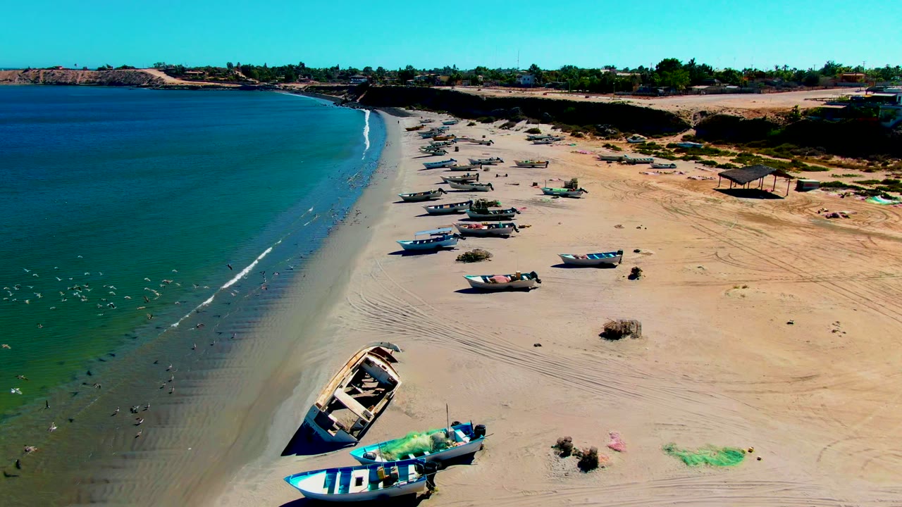 Aerial frame from San Juanico, Baja California Sur showing fishing boats lined along pale sand beside green shoreline water, with no visible people.