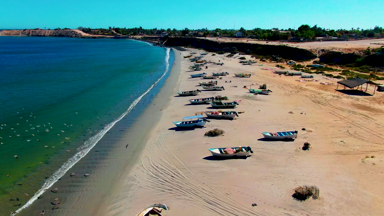 San Juanico aerial frame with multiple fishing boats arranged up the beach, turquoise shallows, darker blue water offshore, and no visible people.