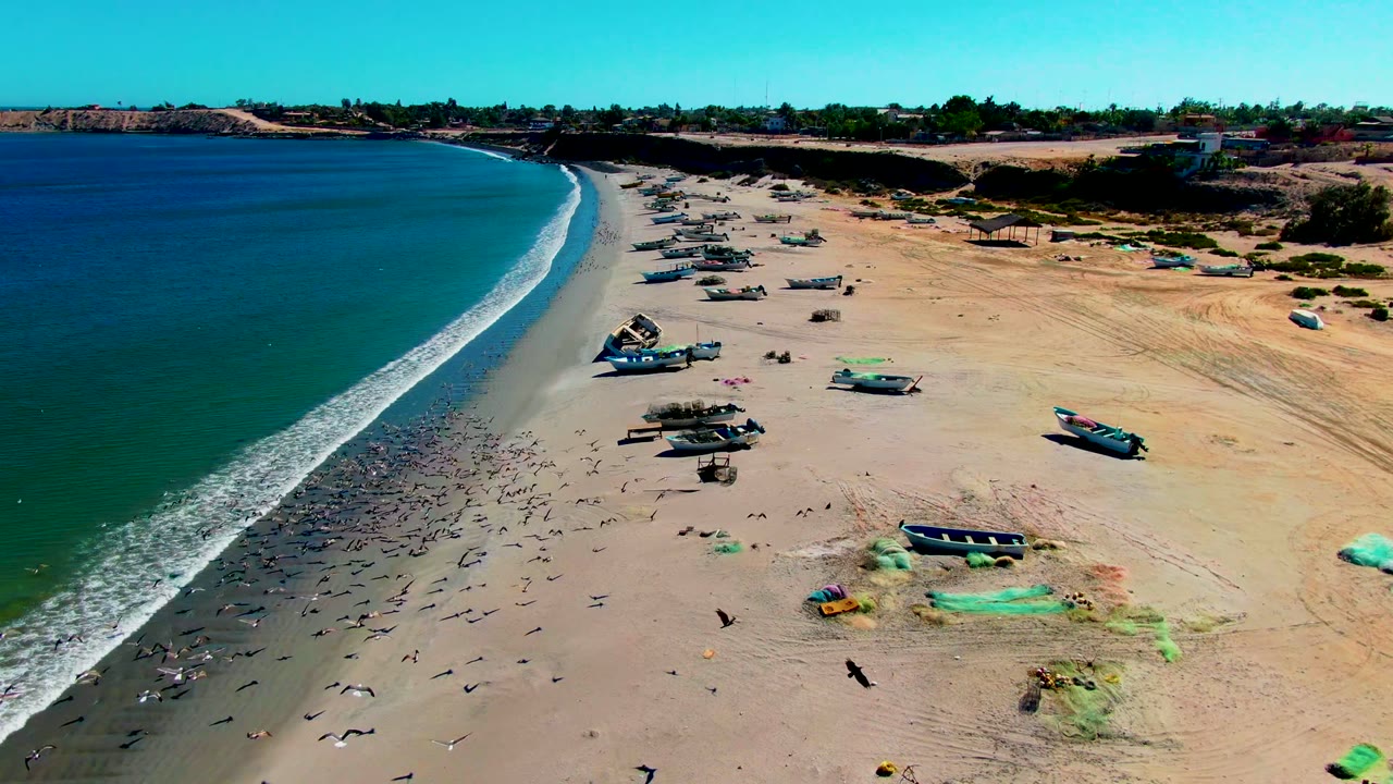 Aerial frame over San Juanico, Baja California Sur showing a curved sandy beach, blue-green ocean at left, several beached fishing boats, shoreline foam, and small people visible near the sand.
