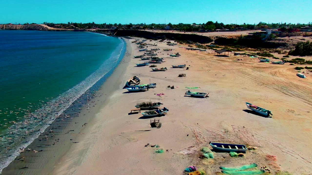 San Juanico beach appears from above with a long diagonal waterline, multiple small fishing boats on pale sand, darker wet shore near the surf, sparse beach gear, and small people visible in the wide frame.