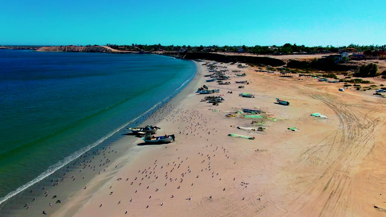 Aerial frame over San Juanico, Baja California Sur showing blue-green water meeting a sandy beach with several fishing boats on shore and no visible people.