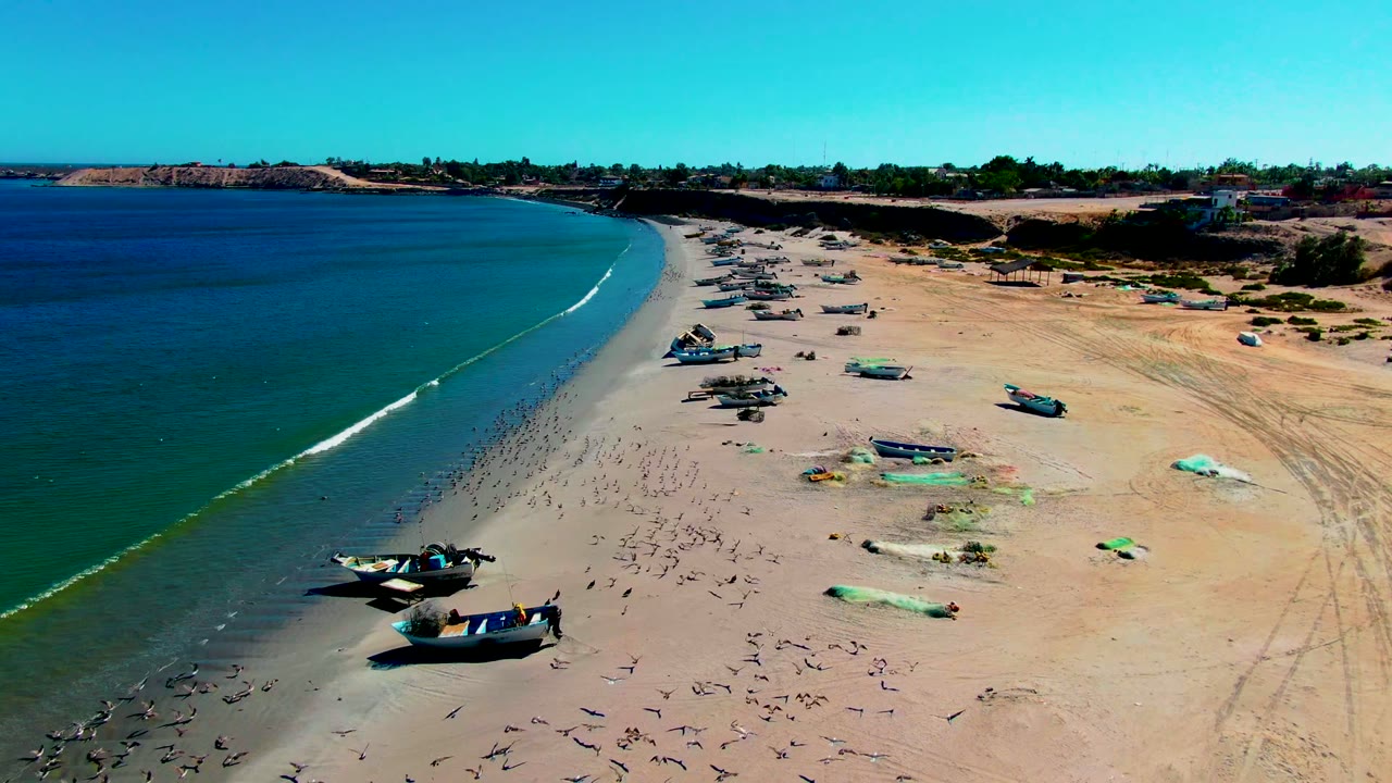 San Juanico shoreline frame with a longer white surf line, multiple boats pulled up on sand, green tarps or nets nearby, and no visible people.
