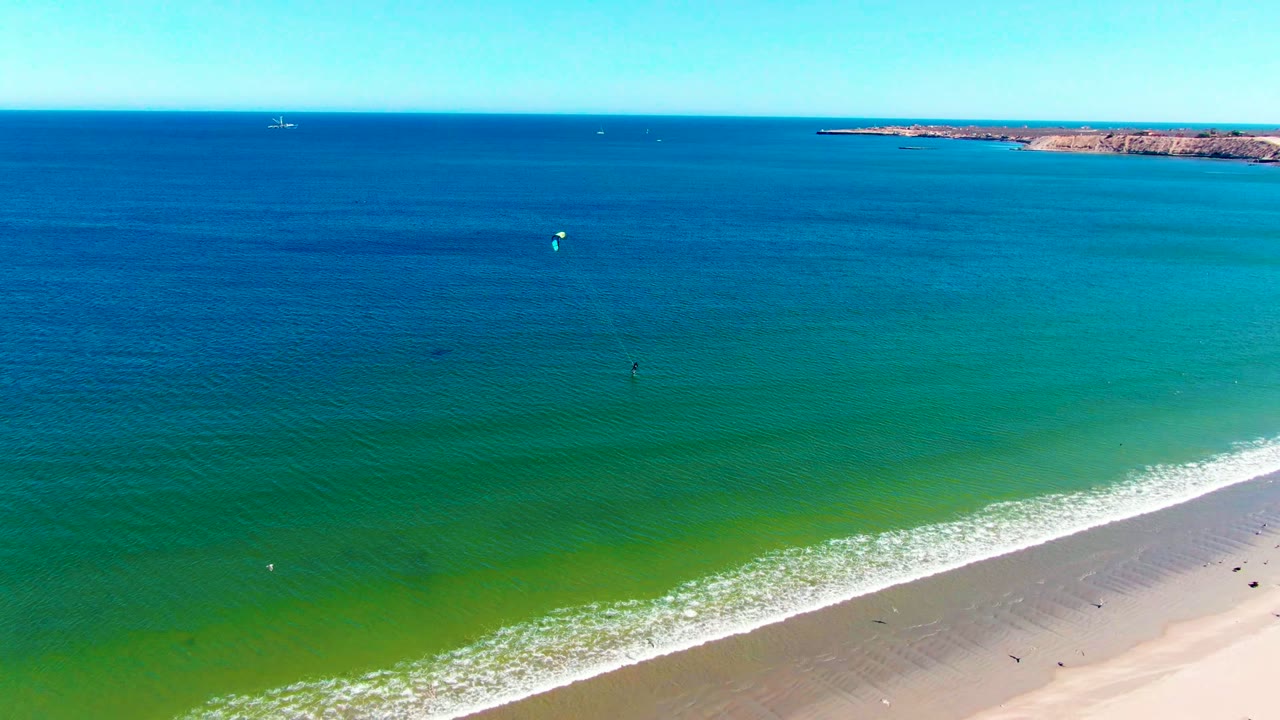Aerial view over San Juanico, Baja California Sur, Mexico, with deep blue water, green shallows, a sandy beach edge, and small people visible in the surf zone.