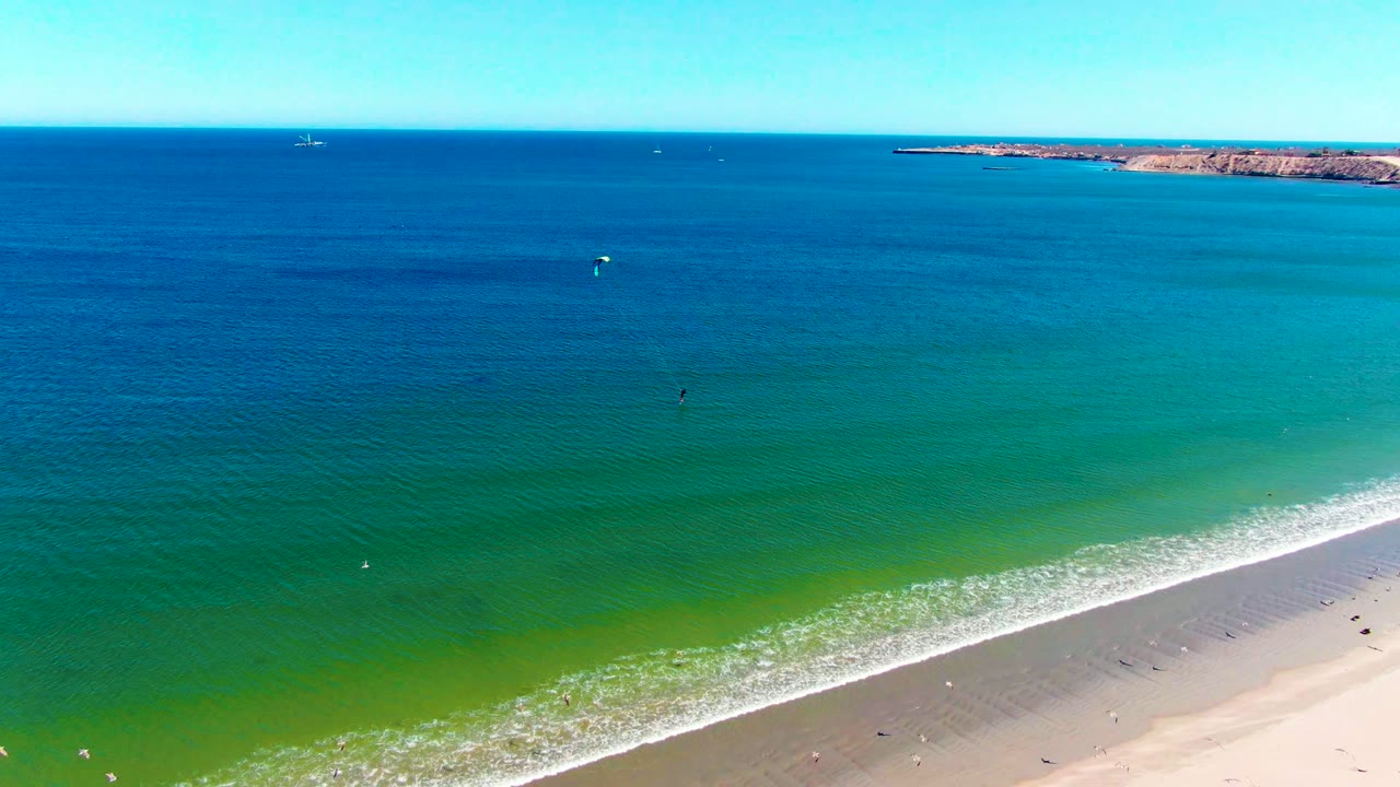 Aerial frame in San Juanico showing turquoise water over the nearshore, a pale sand strip at the lower right, tiny swimmers, and a rocky landform along the horizon.