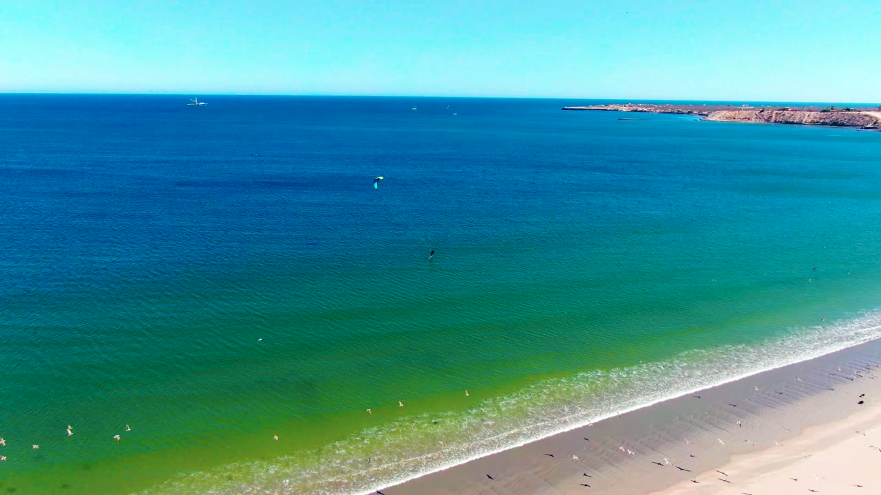 Drone-style aerial view over San Juanico with calm blue ocean, green shallows, a thin white surf line, beachgoers as small figures, and a low rocky point in the background.
