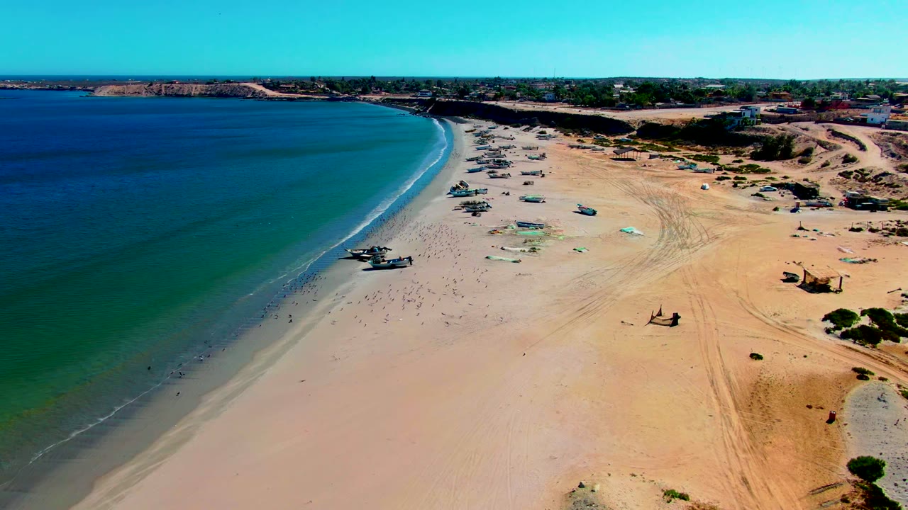 Aerial view of San Juanico beach in Baja California Sur with blue-green water on the left, small boats on the sand, dune tracks to the right, and no visible people.