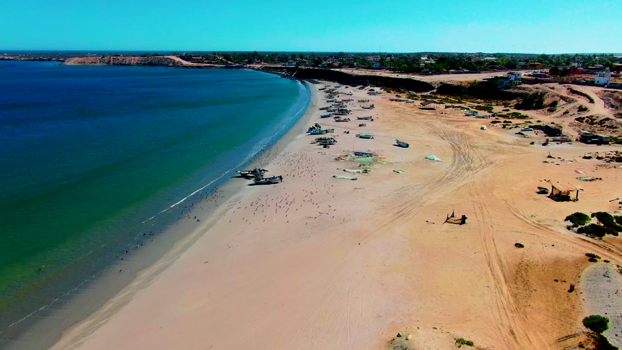Wide aerial coastline with gentle surf and small boats spaced along the sand.