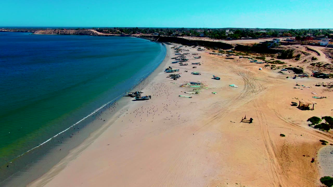 A high coastal view over San Juanico shows the ocean covering the left half of the frame, pale sand with parked boats in the center, dunes on the right, and no visible people.