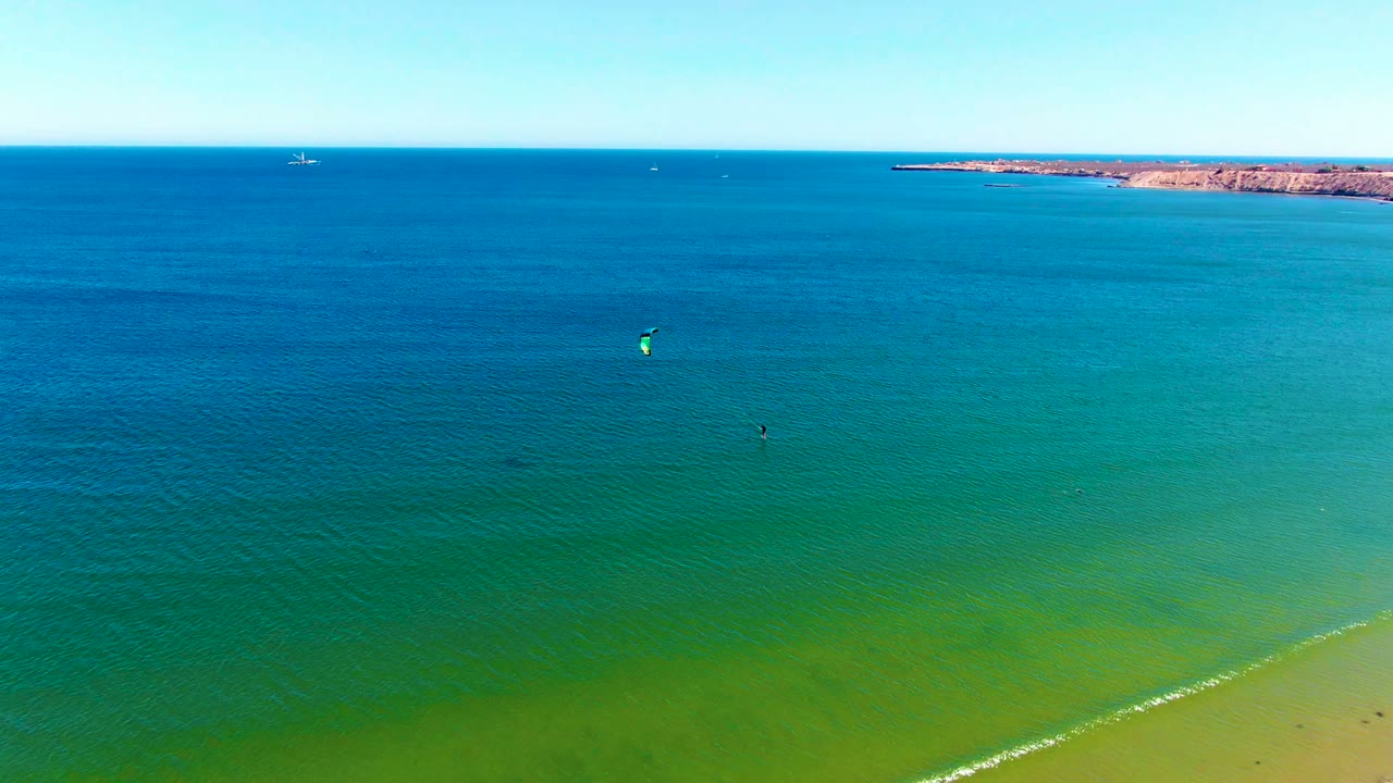 Aerial view in San Juanico, Baja California Sur, with turquoise water filling the frame, a sandy shore at lower right, distant coast on the horizon, and small kite surfers offshore.