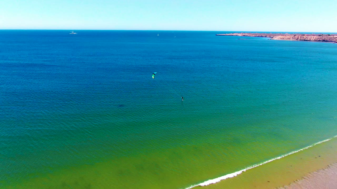 Wide aerial ocean view with a bright kite, distant rider, shoreline foam, and low coast on the horizon.