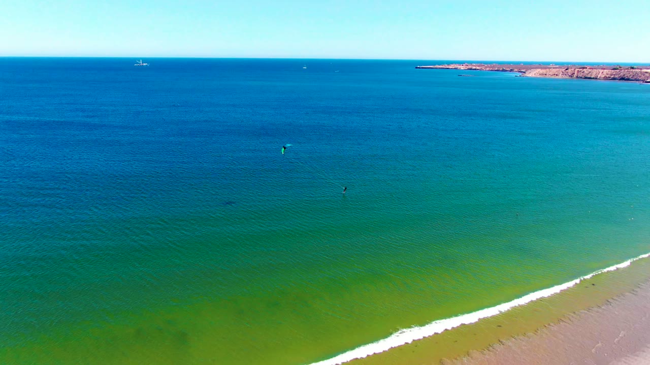 Aerial view in San Juanico with the sandy beach more visible at lower right, a white line of surf, green shallows, blue water, and small kite surfers offshore.