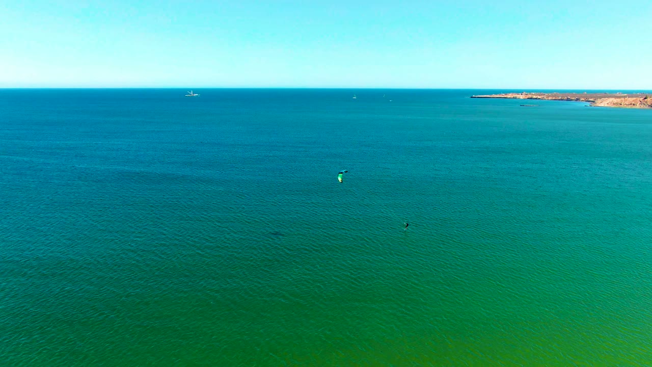 Aerial frame over San Juanico, Baja California Sur ocean water with small kite surfers below, a pale horizon, rocky coast at right, and no visible people.