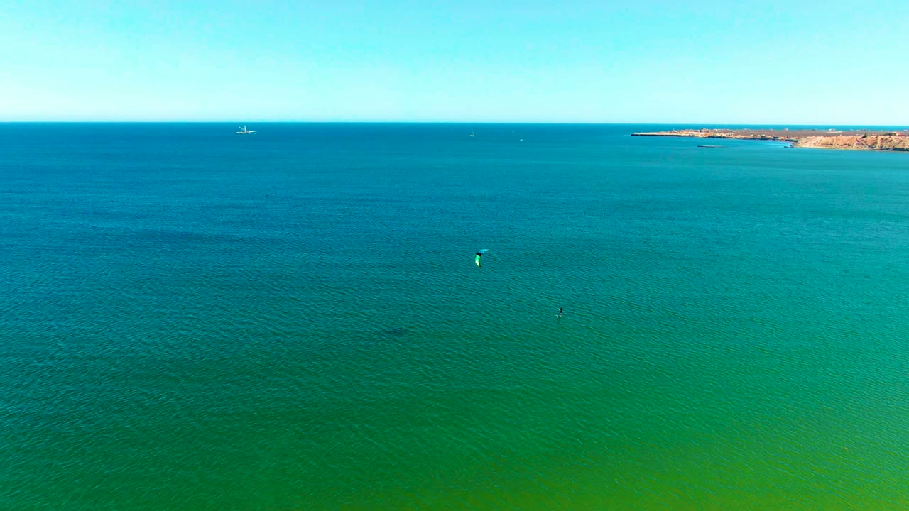 San Juanico aerial frame showing layered blue and green water, tiny kite surfing activity in the midground, rocky coast near the horizon, and no visible people.