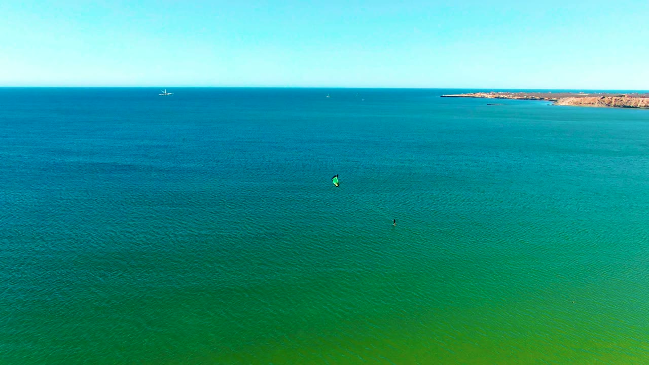 Wide sea horizon with small kite surfers and a rocky point in the background.