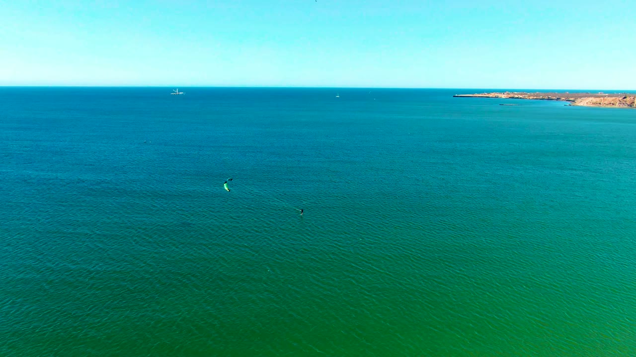 Wide aerial frame over San Juanico ocean water, with two small kite surfers spread across the blue-green surface, a rocky coastline at the horizon, and no visible people identifiable.