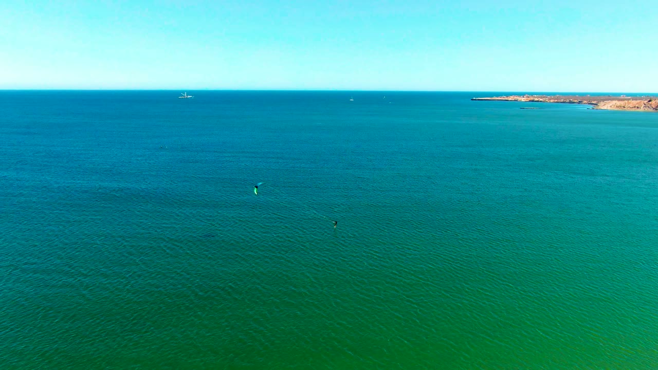 Aerial ocean view with two kite surfers below a clear sky and a low rocky shore in the distance.
