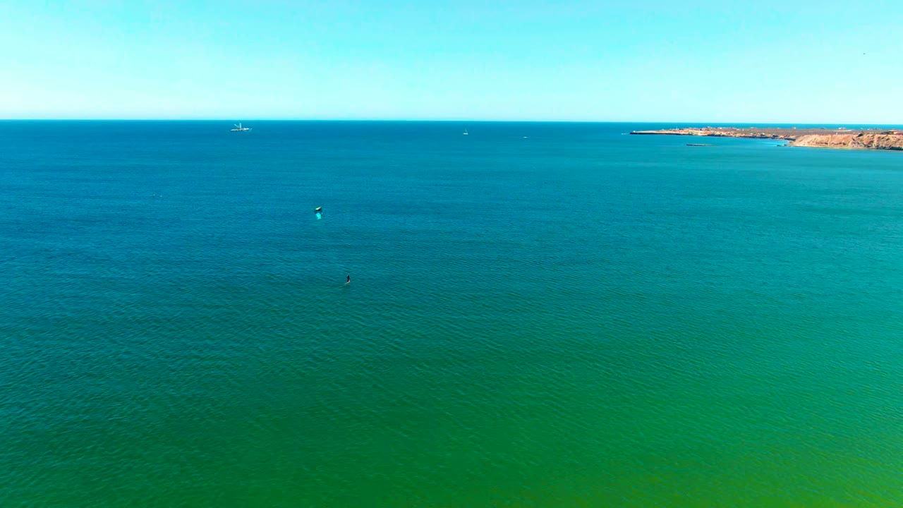 Wide aerial frame over San Juanico turquoise ocean with small kitesurfers scattered across the water and a tan coastline on the horizon.