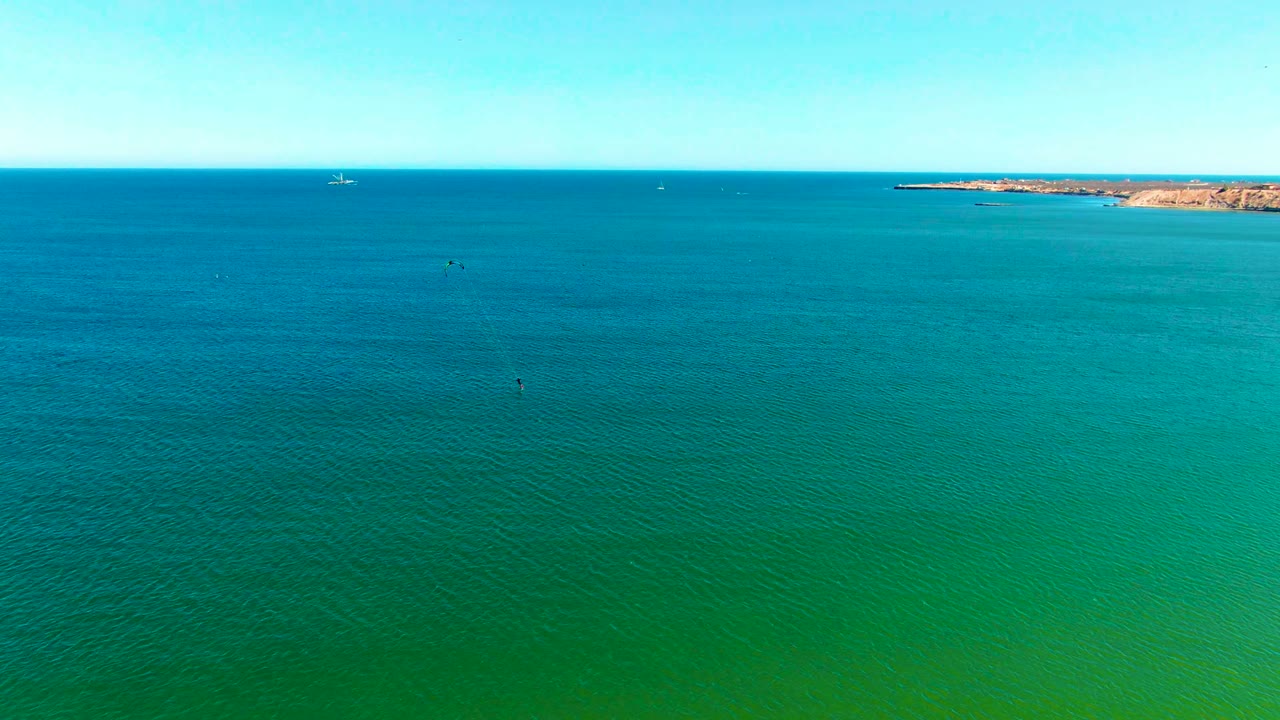 A broad San Juanico seascape shows turquoise water below a clear sky, with distant kitesurfers and a tan headland along the horizon.