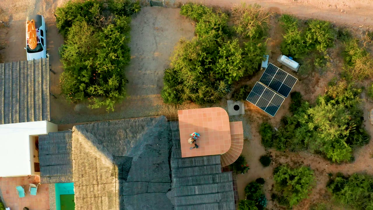 Drone view over the thatched roof house, patio, solar panels, trees, and dirt paths.