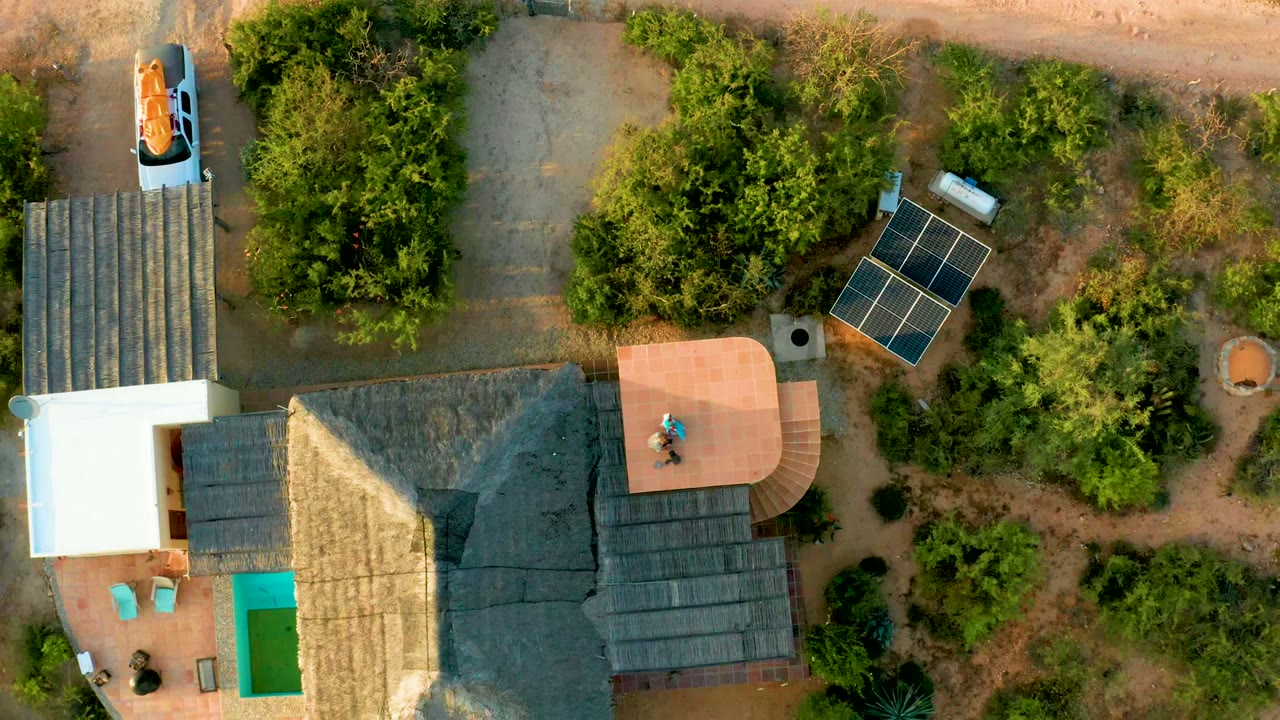 Overhead frame in Cabo Pulmo showing a visible person on the curved patio, the thatched house below, solar panels to the right, trees around the property, and dirt paths above.