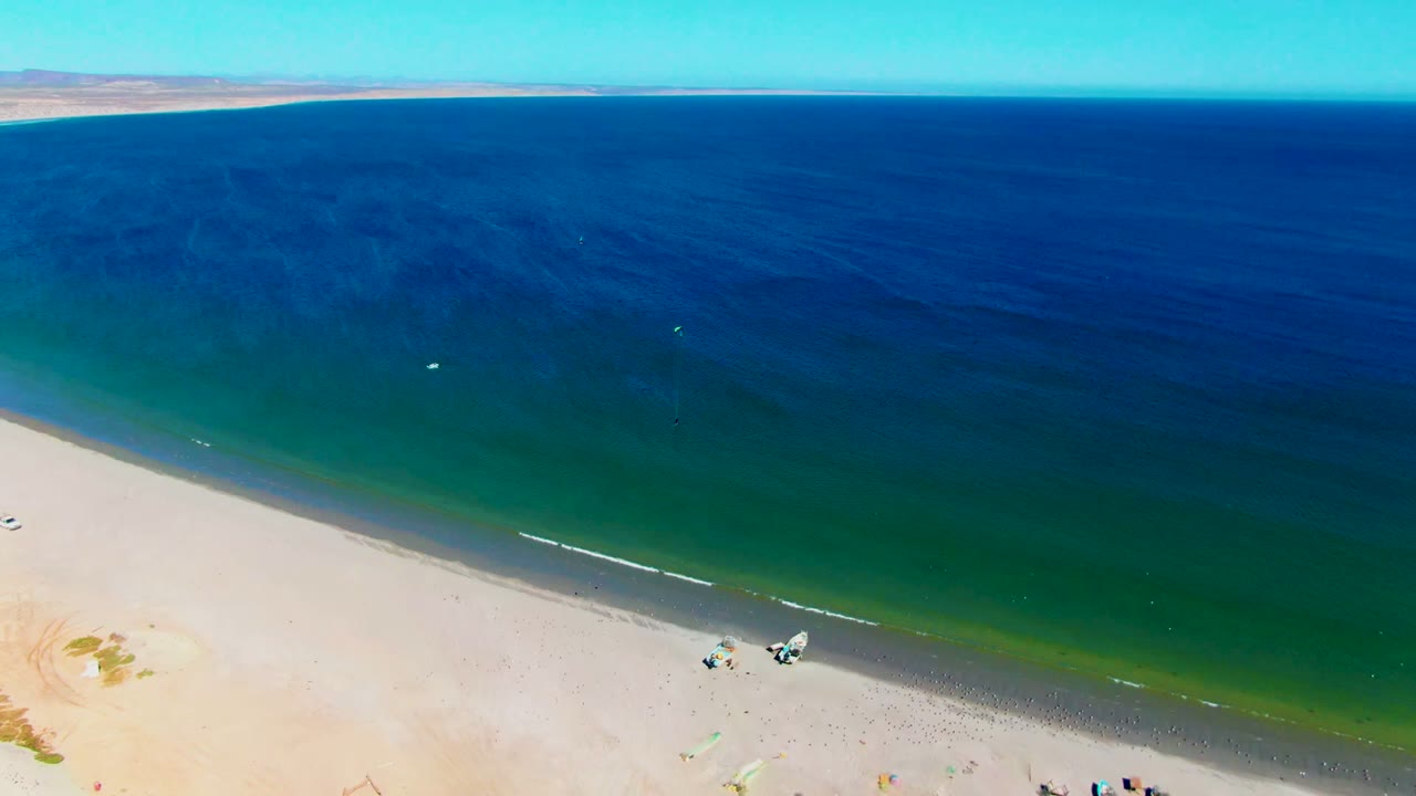 Aerial view over San Juanico beach, with blue-green water, a thin surf edge, pale sand, distant coastline, and no identifiable people visible.