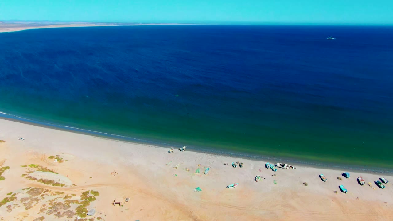 Aerial frame over San Juanico, Baja California Sur, with pale sand at the lower edge, small boats near shore, tiny beachgoers, and deep blue ocean beyond.