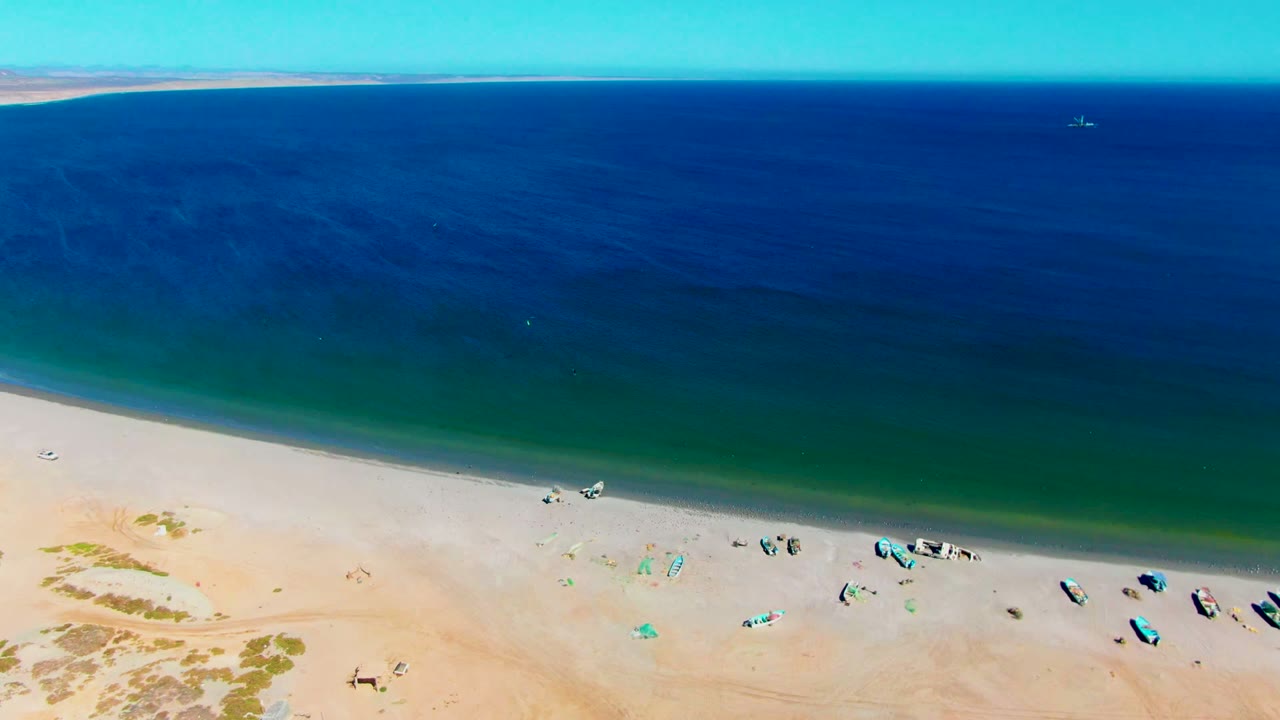 San Juanico beach appears from above with a line of small boats, faint beachgoers, pale sand, and layered blue-to-green ocean stretching toward the horizon.