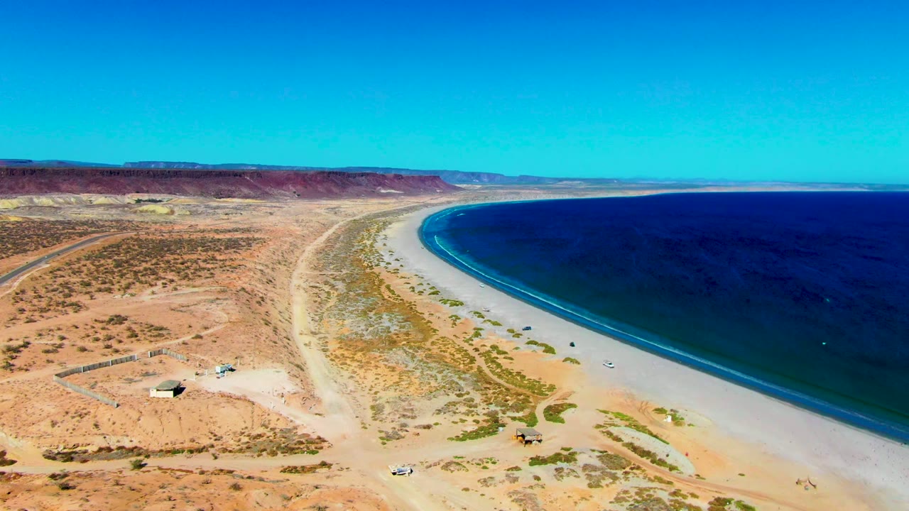 Aerial frame over San Juanico, Baja California Sur, showing desert flats, a curving sandy beach, deep blue ocean, dirt roads, and no visible people.