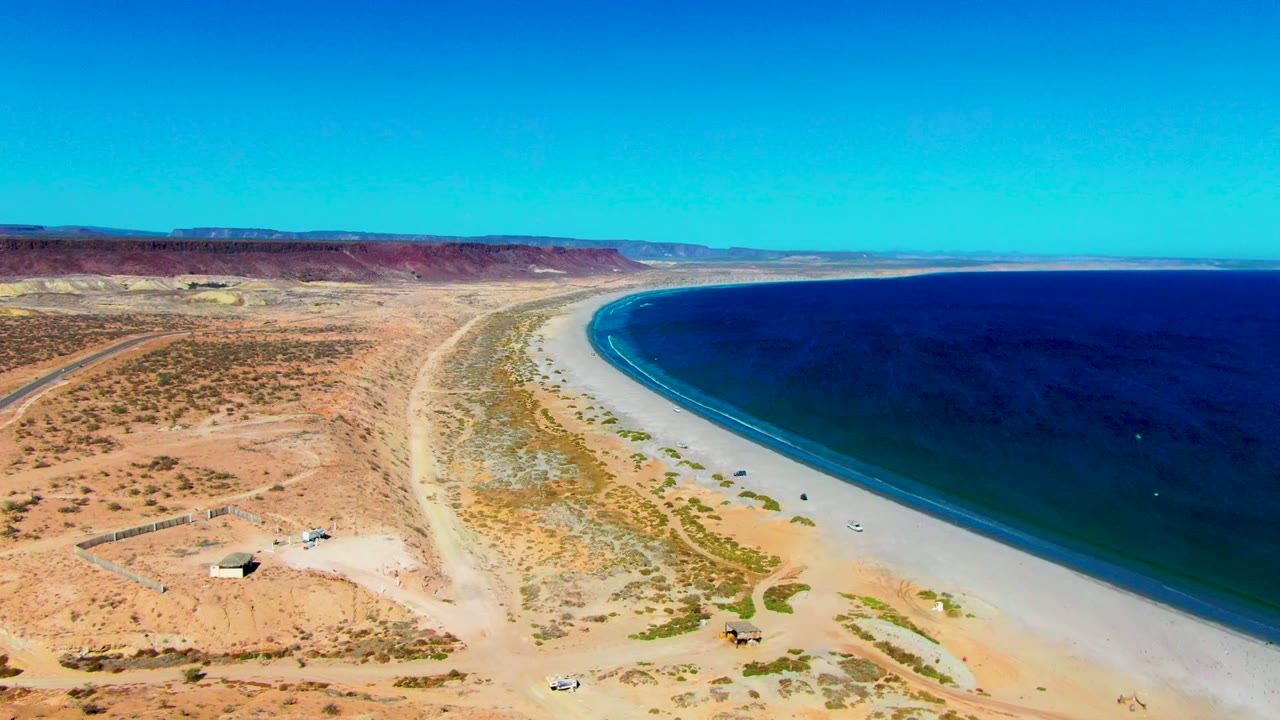 Curving beach bordered by dry desert terrain and dark blue water.
