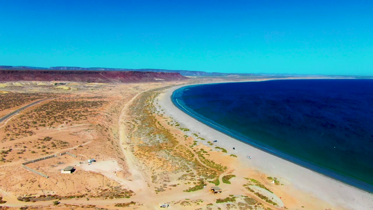 Wide aerial frame over San Juanico, Baja California Sur, showing reddish desert, pale beach, a dark ocean band, faint wave lines, and no visible people.