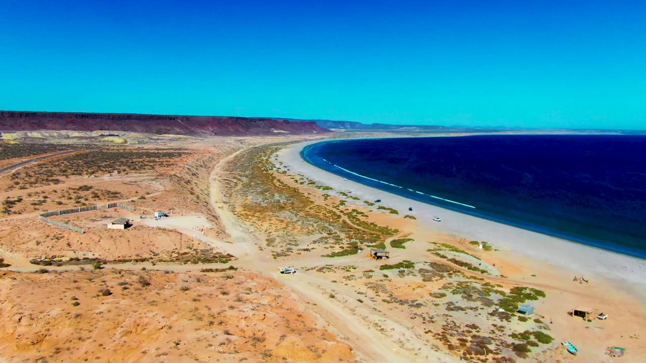 Aerial frame of San Juanico, Baja California Sur showing tan desert scrub, pale beach, curved blue shoreline, white surf, and no visible people.