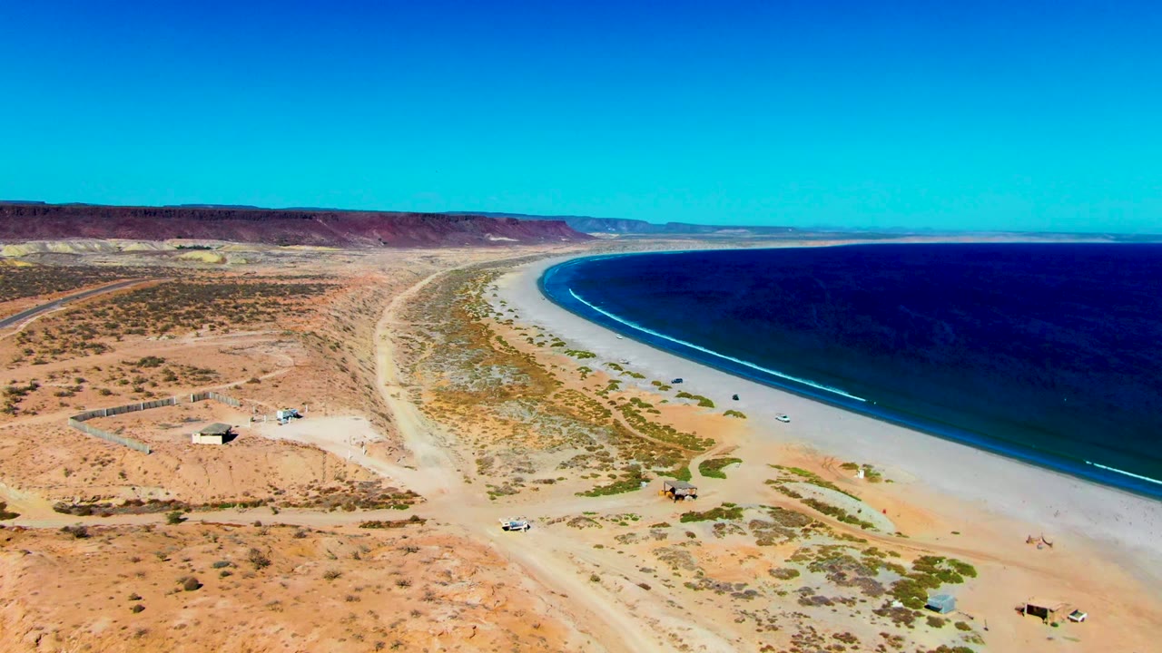 Aerial view in San Juanico showing the long pale beach curving beside dark blue water, with desert scrub and small ground features inland, no visible people.